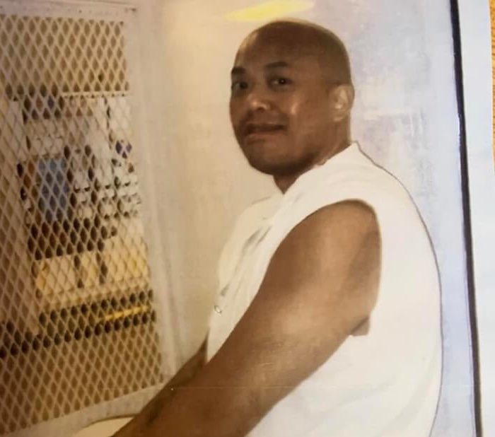 Kosoul Chanthakoummane, a bald Laotian man, sits inside a viewing window in a prison, wearing a sleeveless white top and white pants.
