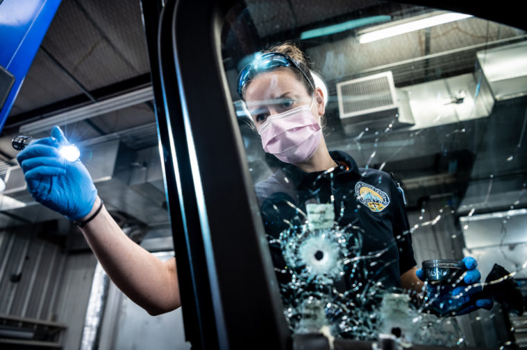 A scientist in a Houston Forensic Science Center uniform uses a flashlight and forensic test kit to study a bullet hole in a vehicle's window.