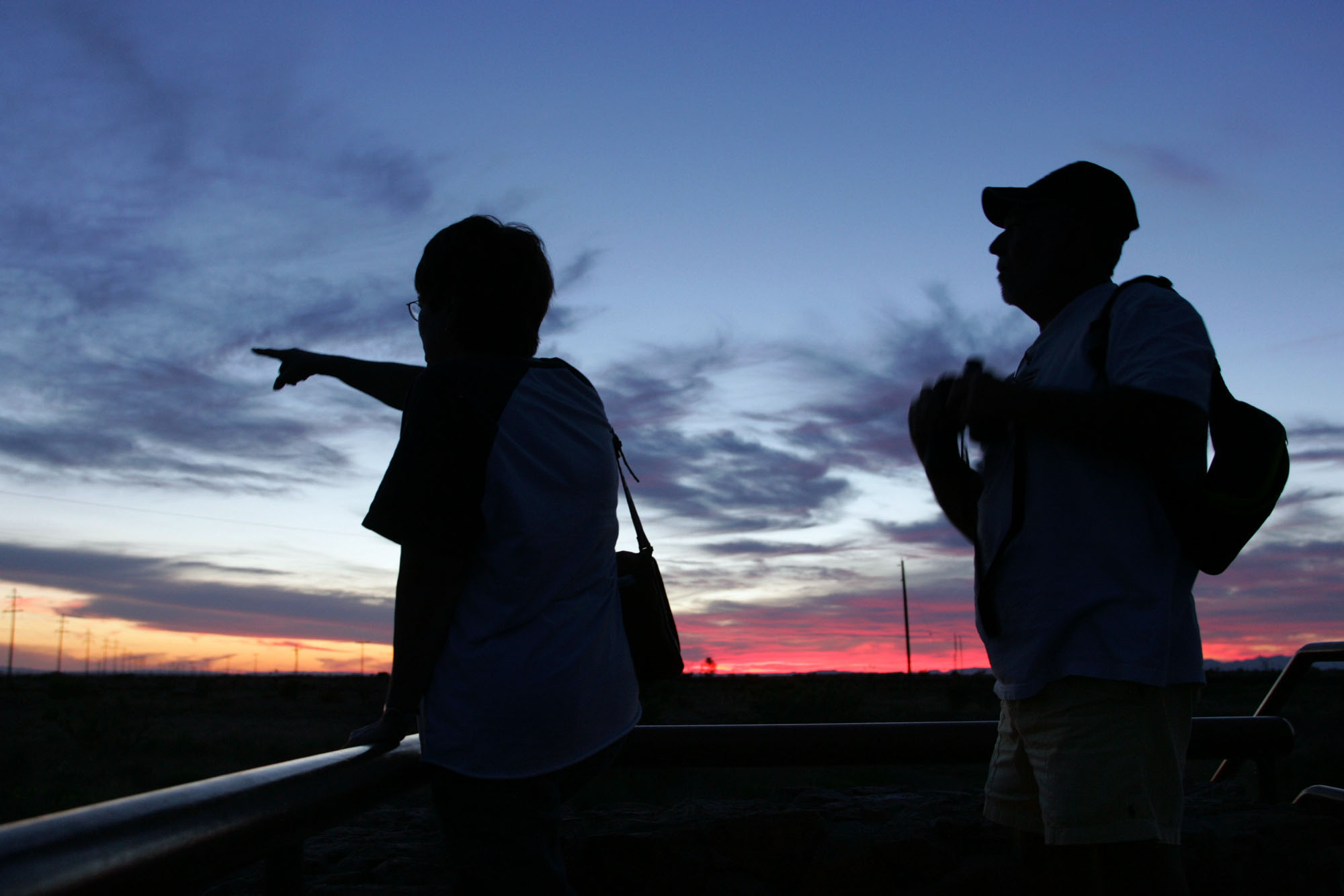 Fern Teems, left, points to what she believes may be one of the mystery lights near Marfa, Texas, as her husband James Teems looks on. Both are silhouetted against a blue, purple, and vibrant red sunset sky.