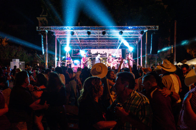 A large rock concert crowd, seen from behind looking up at the brightly lit stage where a band is performing. Many in the crowd are wearing cowboy hats.