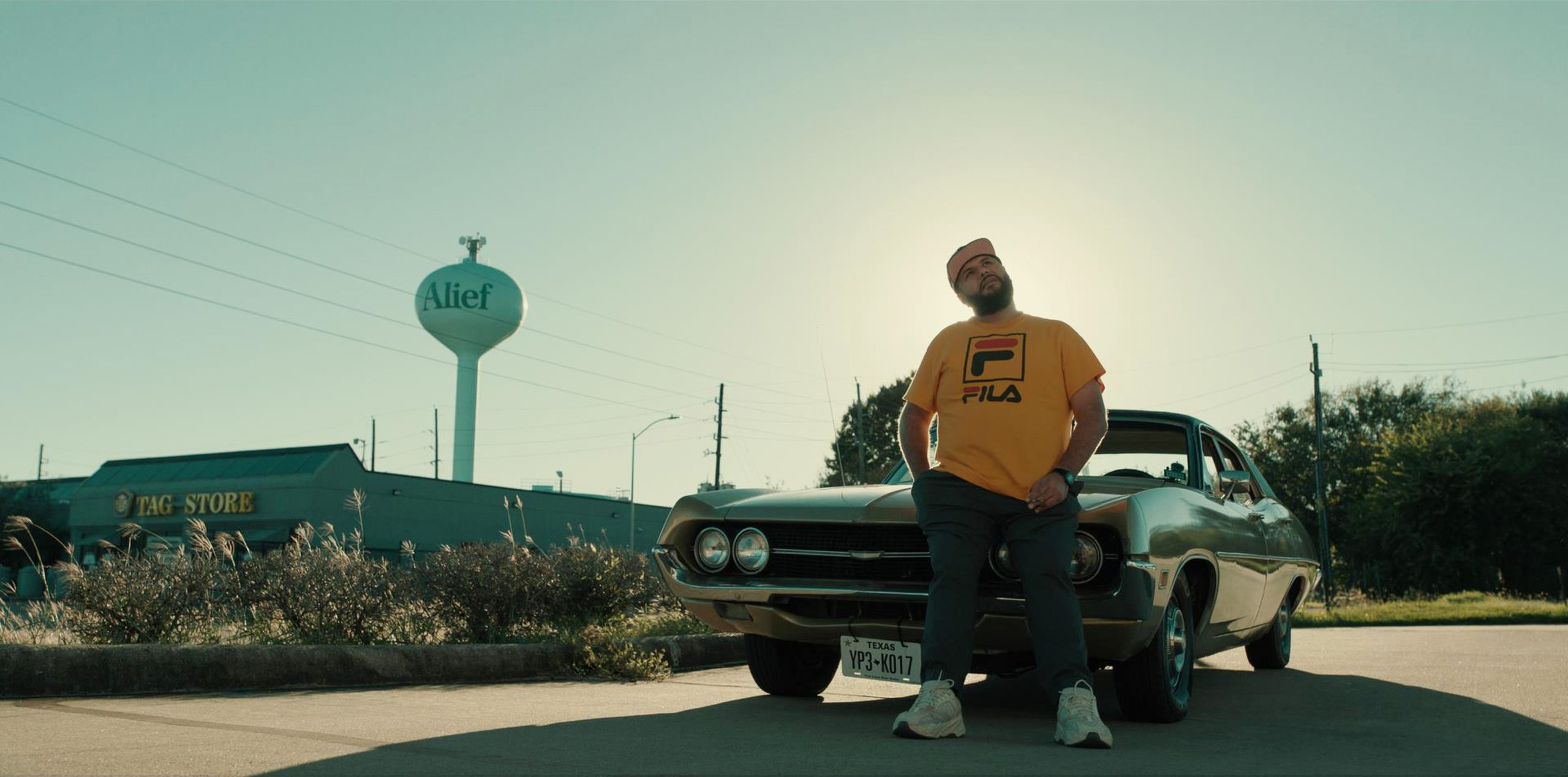 Mo, a Palestinian American with a thick short beard, leans against a Dodge challenger, near a Houston suburban strip mall with a water tower in the background. He's wearing a white baseball cap, a yellow FILA shirt and baggy blue jeans.