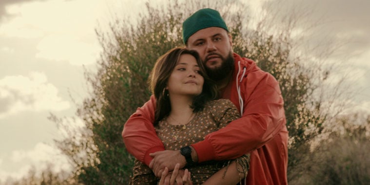 Mo Amer embraces Teresa Ruiz, wrapping his arms around her from behind, artfully posed with a bush and the cloudy sky behind.