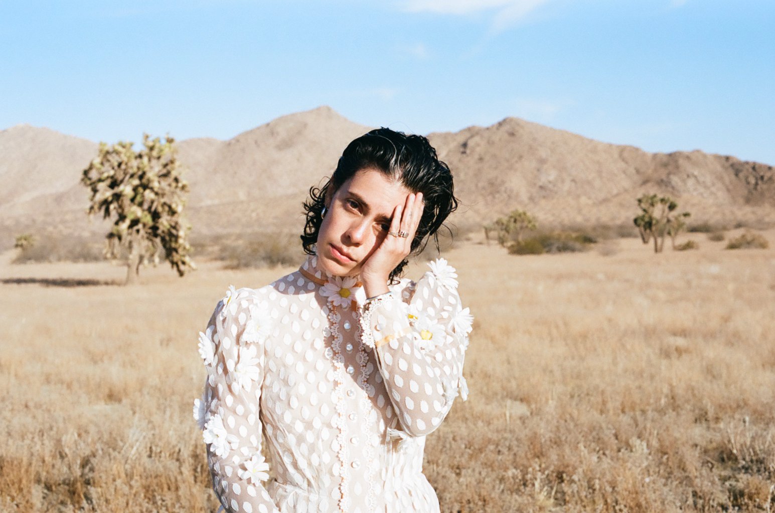 Roberta Colindrez, a Latinx woman poses in a prairie setting, with mountains and desert scrub plants and succulents behind her. She's wearing an old-fashioned white lace dress covered in white teardrop shapes and frilly buttons down the front, and playfully cradling hear face in one hand. She has shoulder length, slightly curly dark hair.