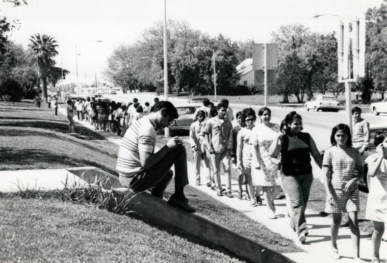 A youg man writes notes sitting on a staircase as protesters march by on a sidewalk in Uvalde in 1970.