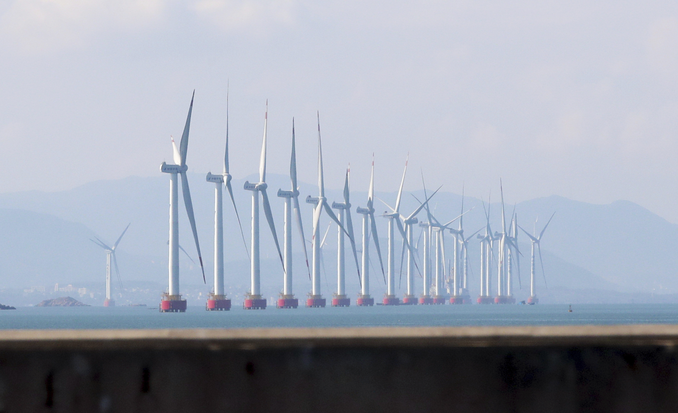 A row of massive power generating windmills can be seen floating on the water offshore.
