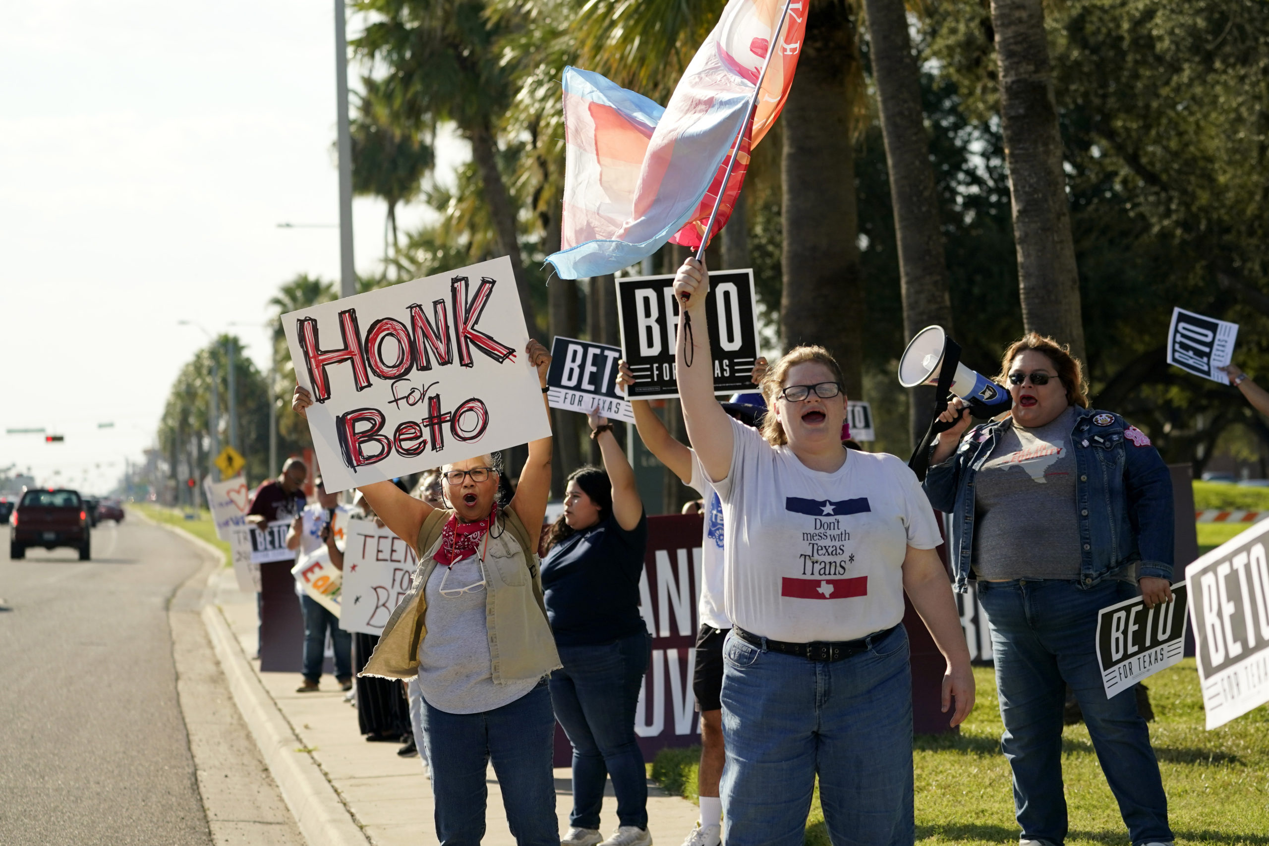 Beto supporters wave signs, flags and hold a banner by the side of a road, while another uses a bullhorn.