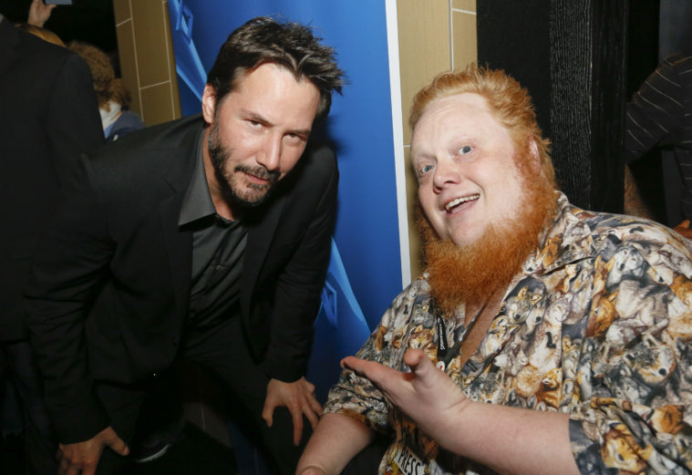 Harry Knowles, a redhead in a very loud Hawaiian shirt, wears an excited expression as he poses with Keanu Reeves, in a suit with no tie.