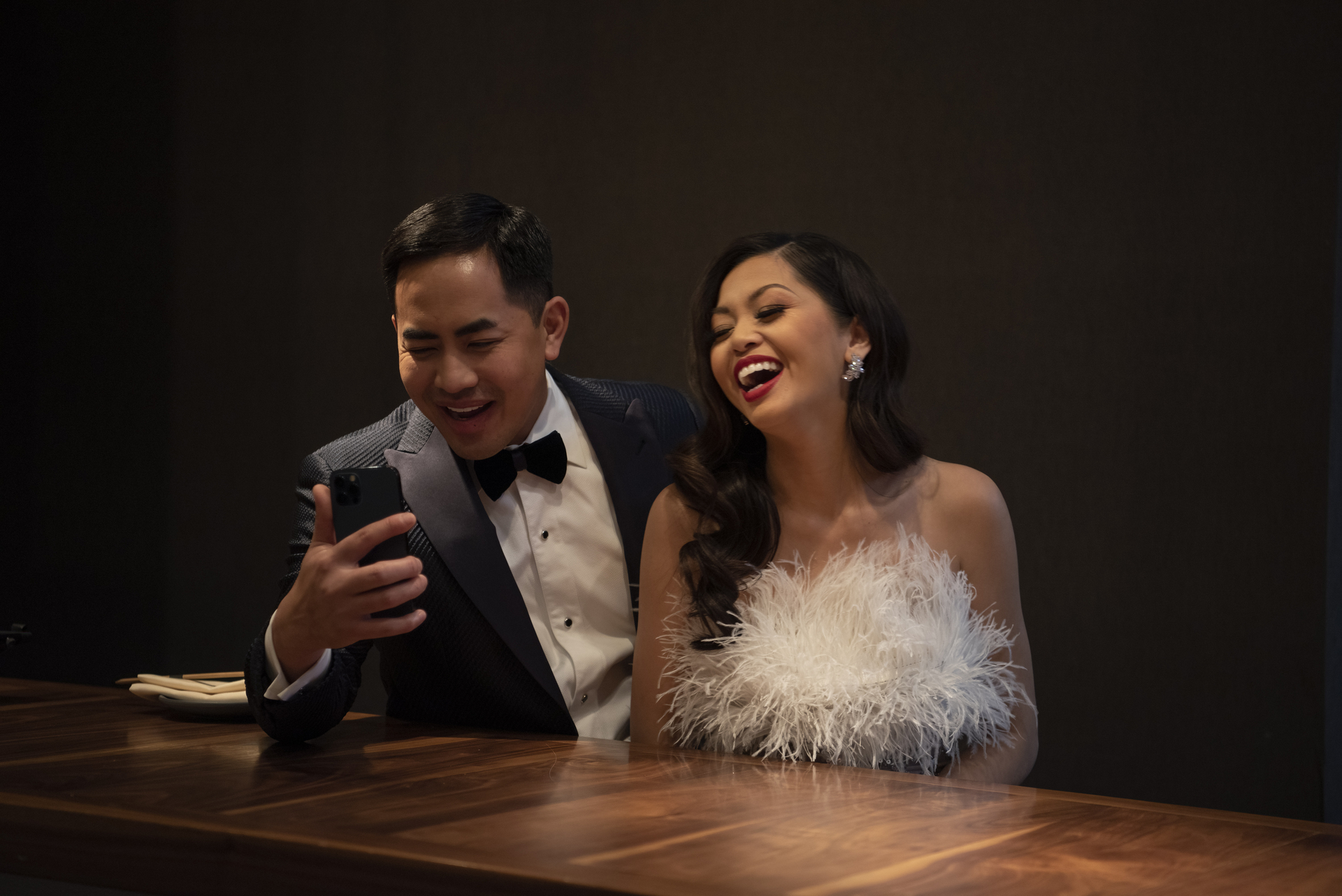 Washington Ho, in a tuxedo with bow tie, and Lesley Ho, in a fluffy white designer dress, are seated and laughing theatrically as they look into a phone as if taking a selfie or on a video call.