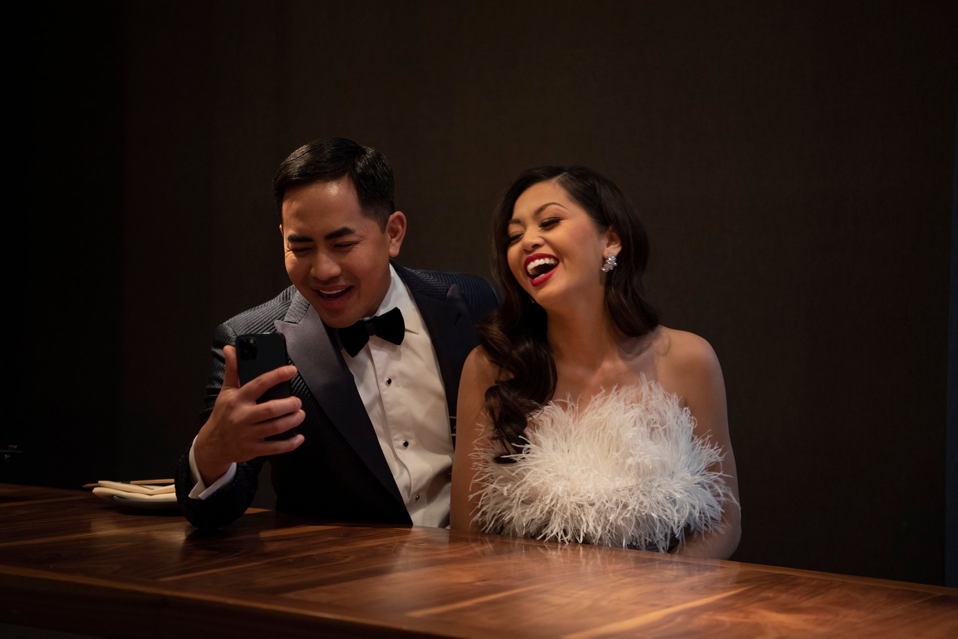 Washington Ho, in a tuxedo with bow tie, and Lesley Ho, in a fluffy white designer dress, are seated and laughing theatrically as they look into a phone as if taking a selfie or on a video call.