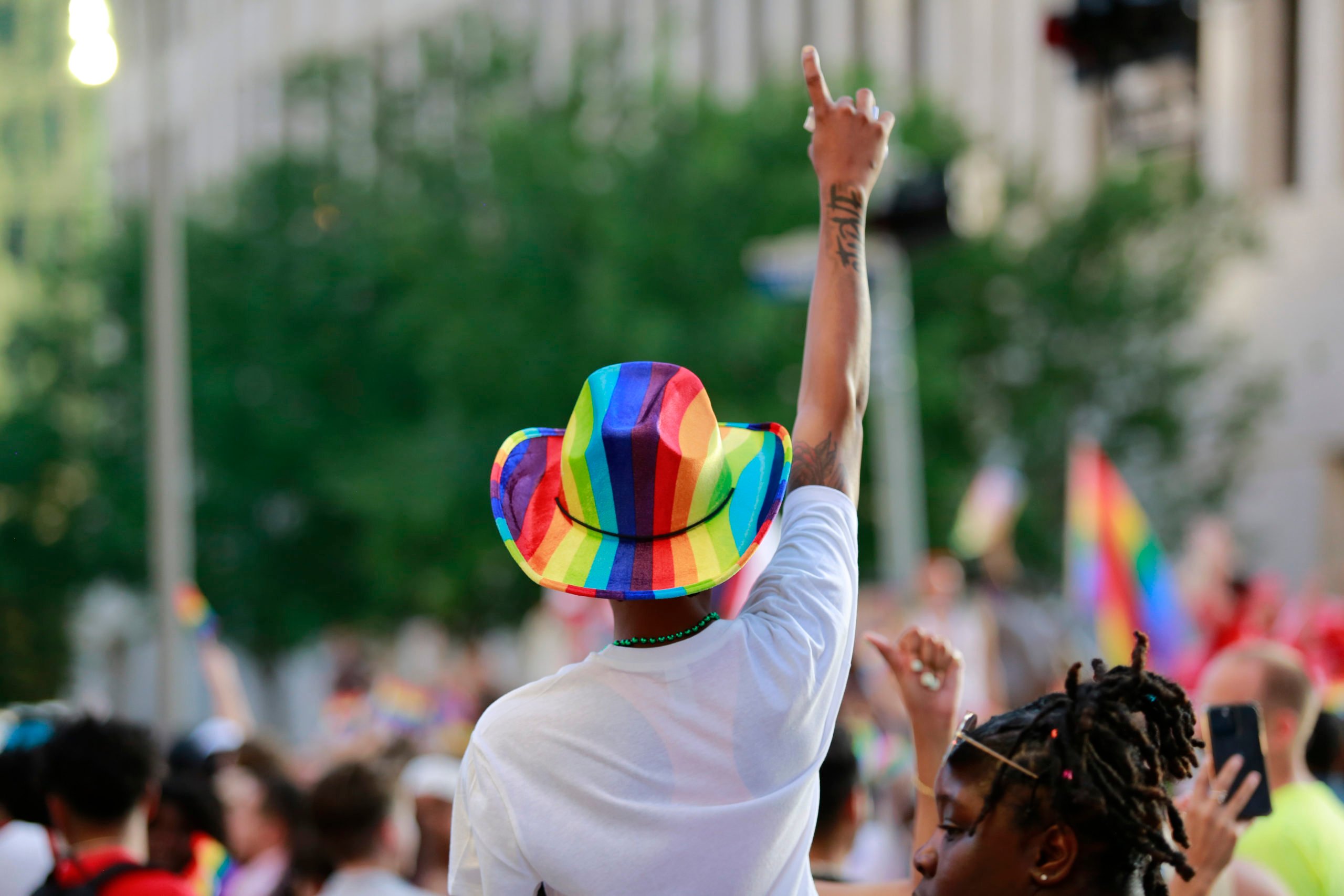 At a Pride Parade in Houston, a marcher in a white tee and pride rainbow cowboy hat salutes with an arm upraised. In the blurry background are more marchers with Pride flags.