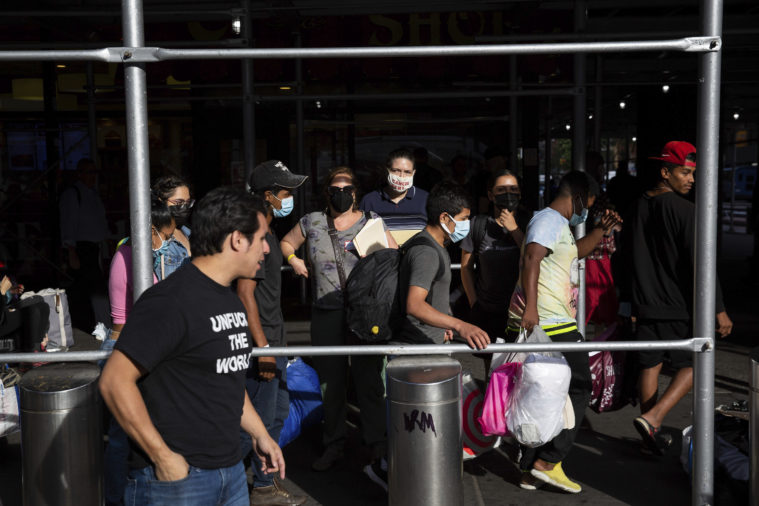 Immigrants line up along a metal barrier after exiting a bus from Texas. One observer or volunteer is wearing a black t-shirt which reads "Unfuck the World."