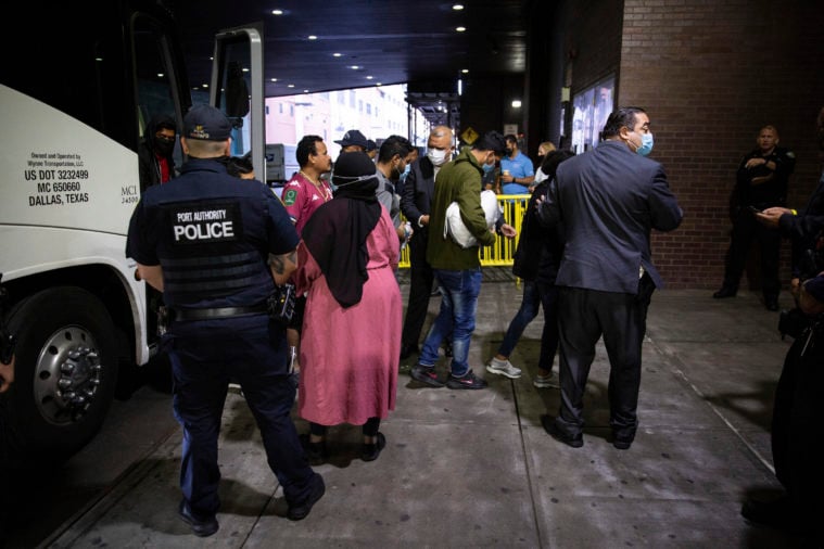This color photo echos the feature photo of the article, showing a modern group of immigrants getting off a bus in New York City after being sent from Texas. one woman at the center of the photo wears a bright pink dress with a black hijab. Port Authority police and city officials are present to help them.