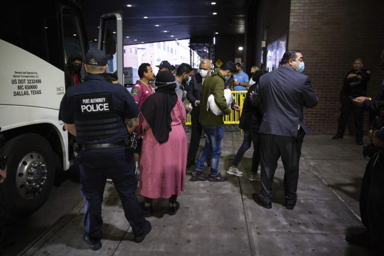 This color photo echos the feature photo of the article, showing a modern group of immigrants getting off a bus in New York City after being sent from Texas. one woman at the center of the photo wears a bright pink dress with a black hijab. Port Authority police and city officials are present to help them.