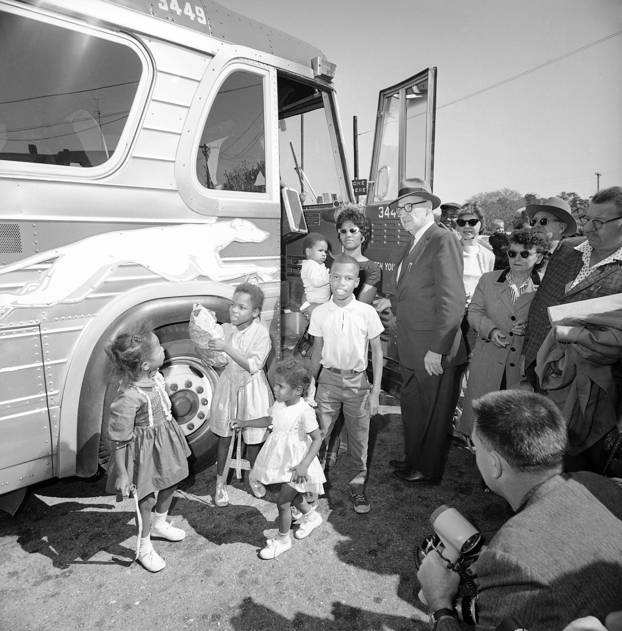 In this historical black and white photo, a group of Black people disembark from a bus, wearing 1960s era clothing.