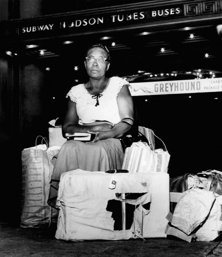 In this black and white historic photo, a Black woman sits atop a pile of luggage, holding a book in her hands. She's wearing cats eye glasses and simple 1960s-era clothing.