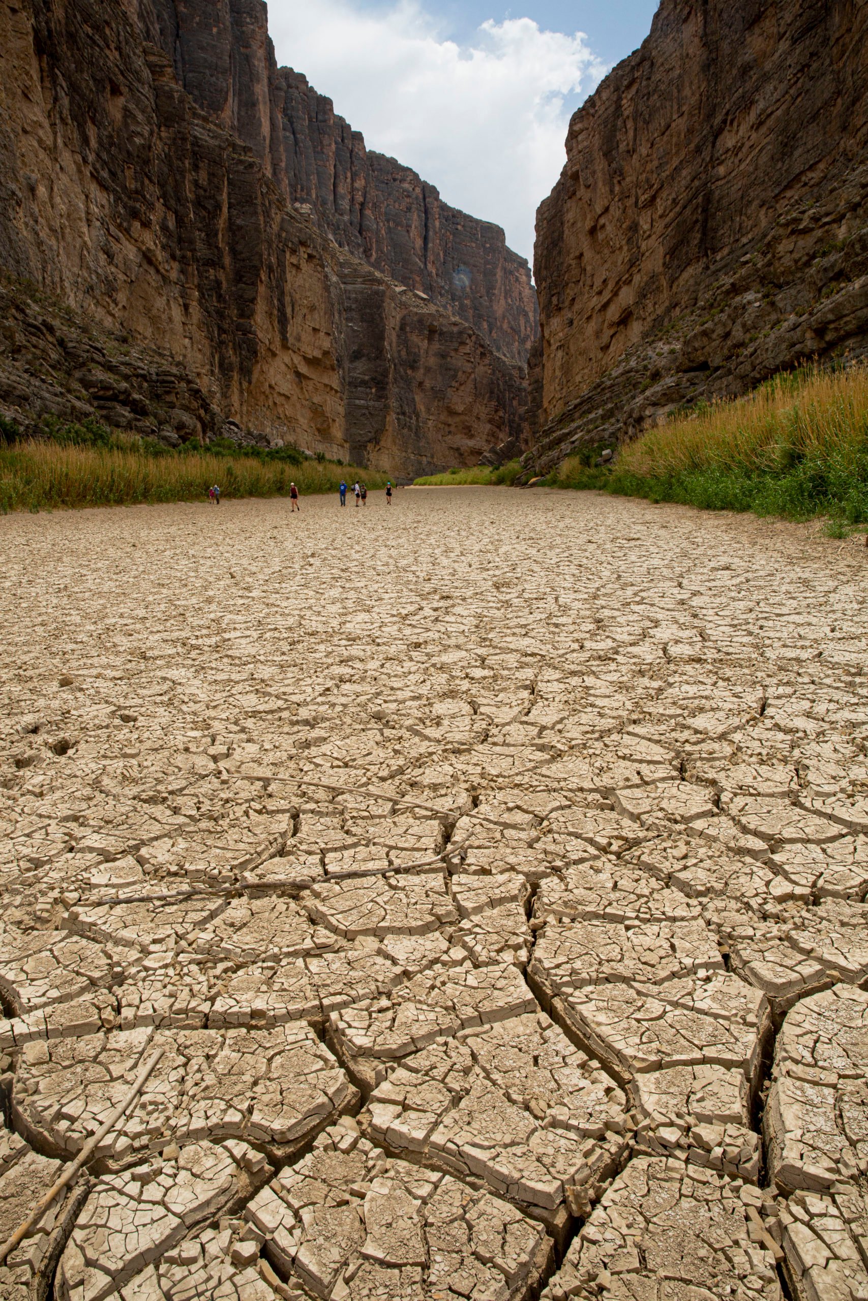 A few solitary figures can be seen walking on the cracked, dry earth where the Rio Grande once ran, lined on either side by tall canyon walls curving into the distance.