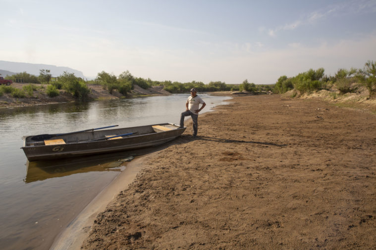 A ferryman stands by his boat, one foot up on the stern, in the dry bed of the Rio Grande.