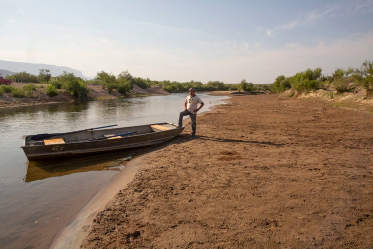 A ferryman stands by his boat, one foot up on the stern, in the dry bed of the Rio Grande.