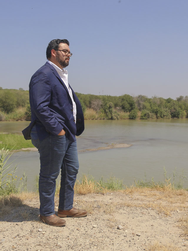 A Latino man in a suit stands with his hands in his pockets, looking out over a river with low water levels.