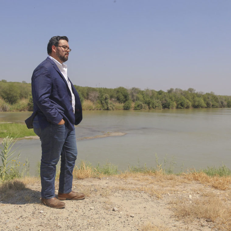 A Latino man in a suit stands with his hands in his pockets, looking out over a river with low water levels.