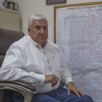 A Latino man with white hair in a button down shirt sits in a chair in front of a water map.