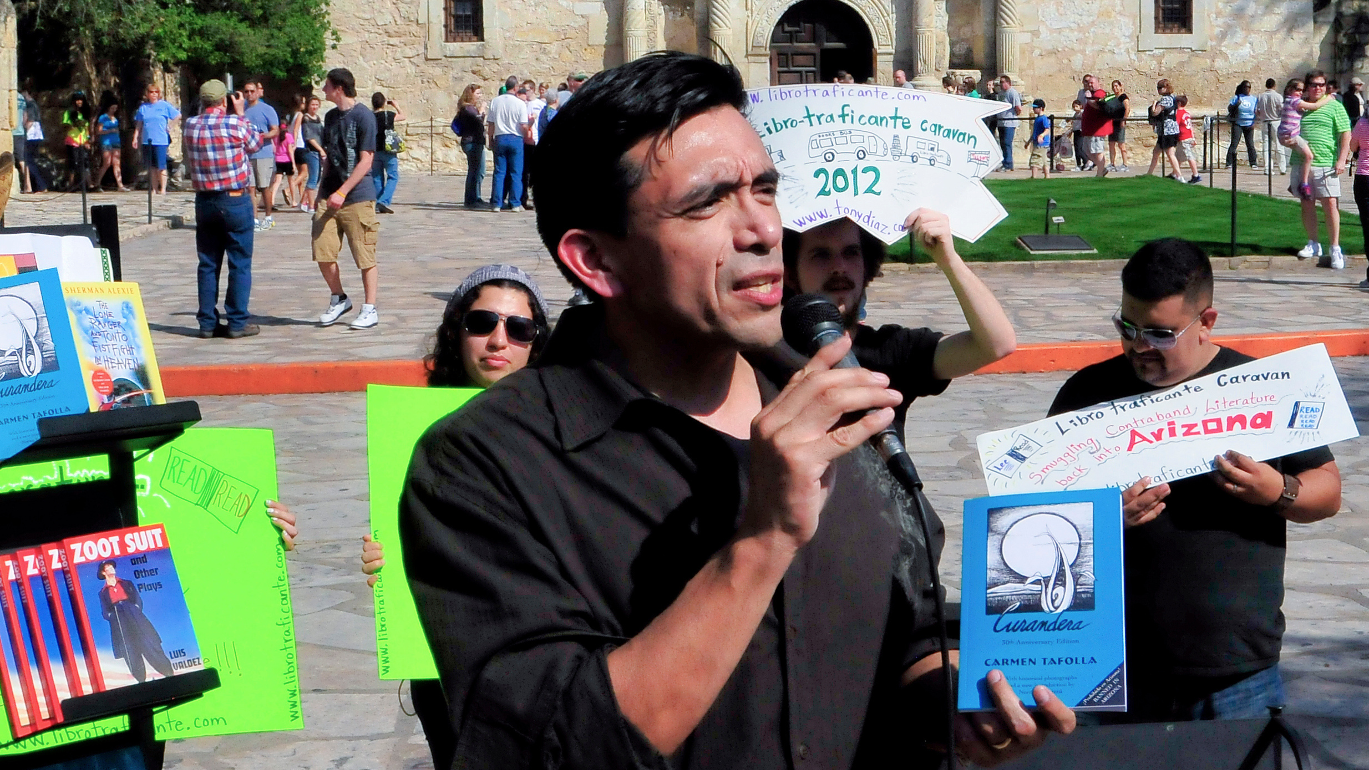 Tony Diaz, a Chicano man with short dark hair and wearing a dark shirt, speaks into a microphone at a protest against book bans at the Alamo.