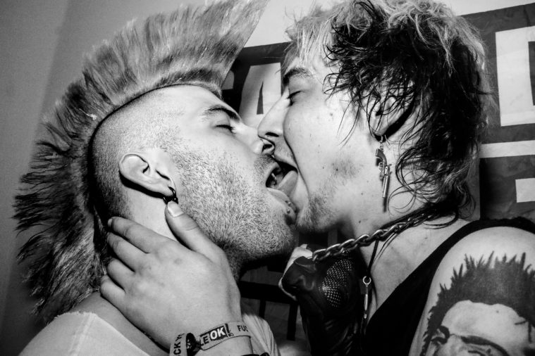 Two masculine looking punks with stubble and fashionable punk outfits, one in a mohawk, make out with lots of tongue.