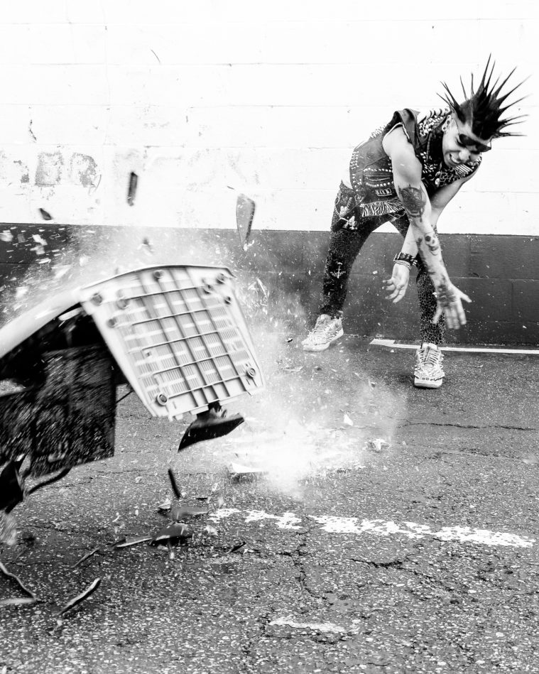 In a black and white action shot, a punk in a mohawk smashes a television onto a cracked pavement street. Glass shatters everywhere.
