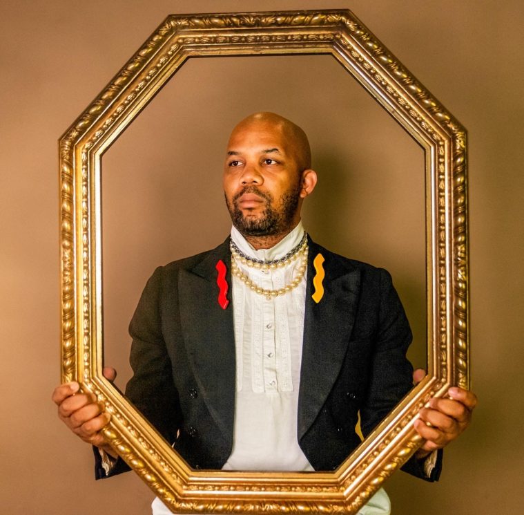 A Black man in a suit jacket and white turtleneck holds a gold picture frame in front of his torso to create a portrait effect.