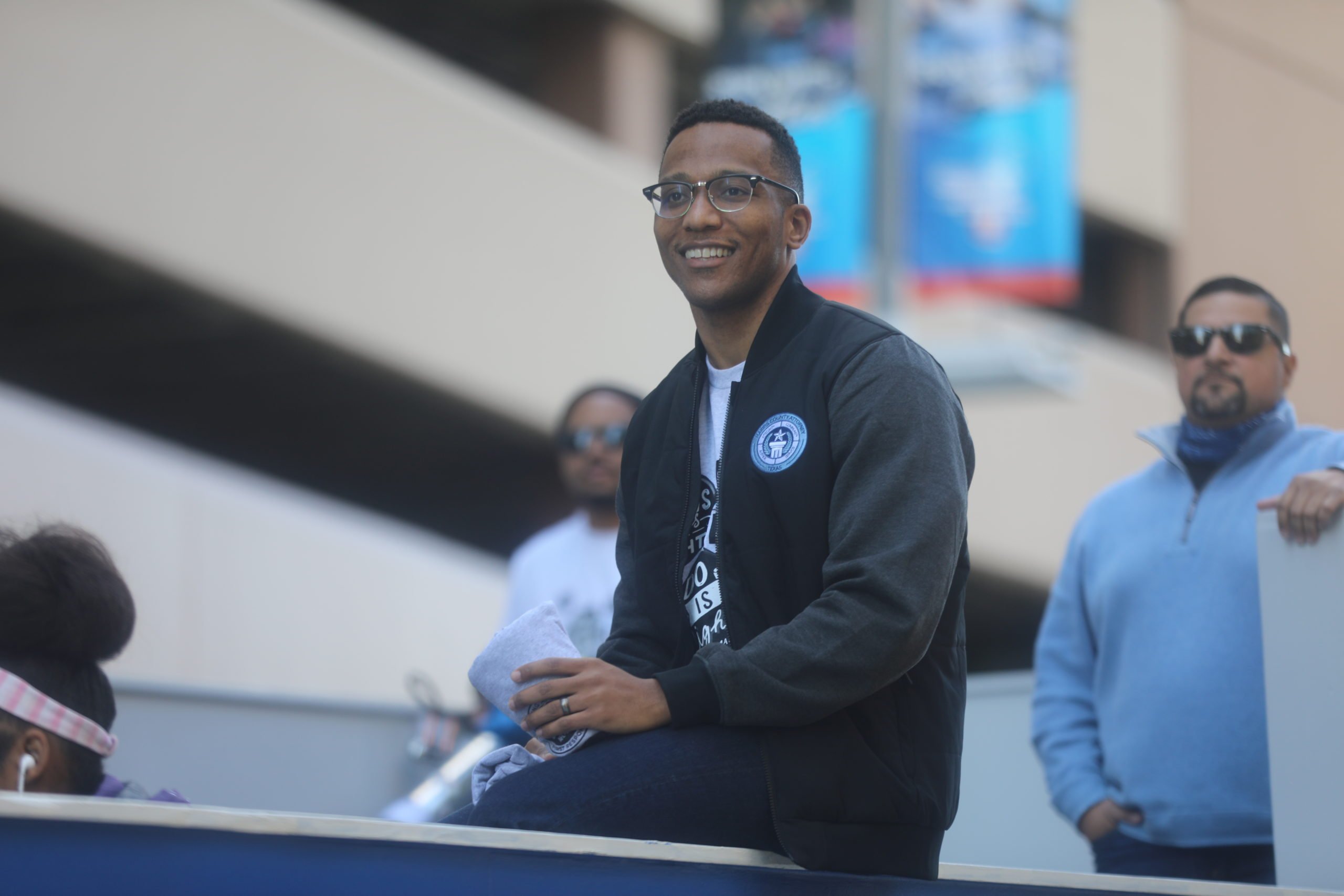 Christian Menefee smiles as he sits on a wall, dressed semi-casually in a Harris County Sweatshirt and his trademark glasses.