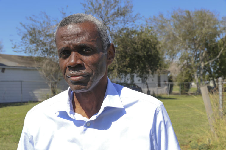 An older Black man with short white hair, dressed in a white button down stands in a back yard, wearing a serious expression.