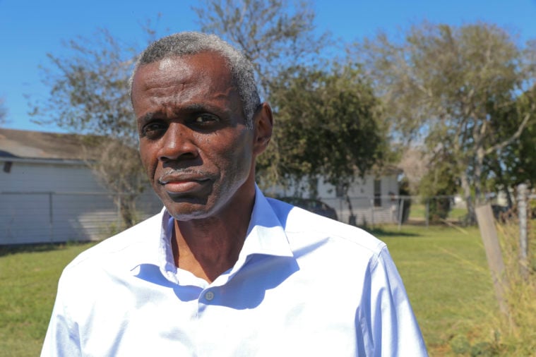 An older Black man with short white hair, dressed in a white button down stands in a back yard, wearing a serious expression.