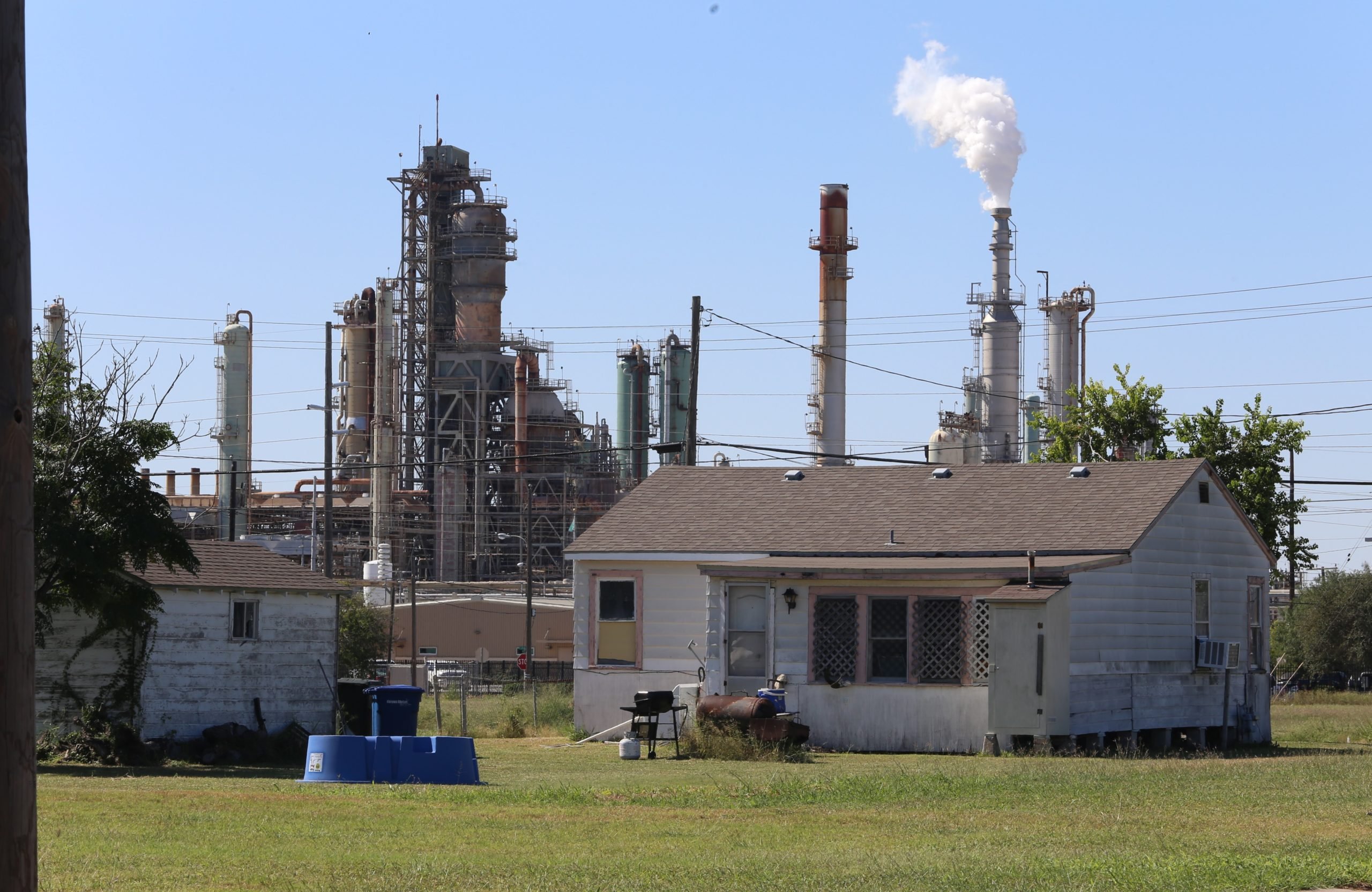 A small house is dwarfed by the refinery behind it, with a smokestack spewing white smoke or steam.