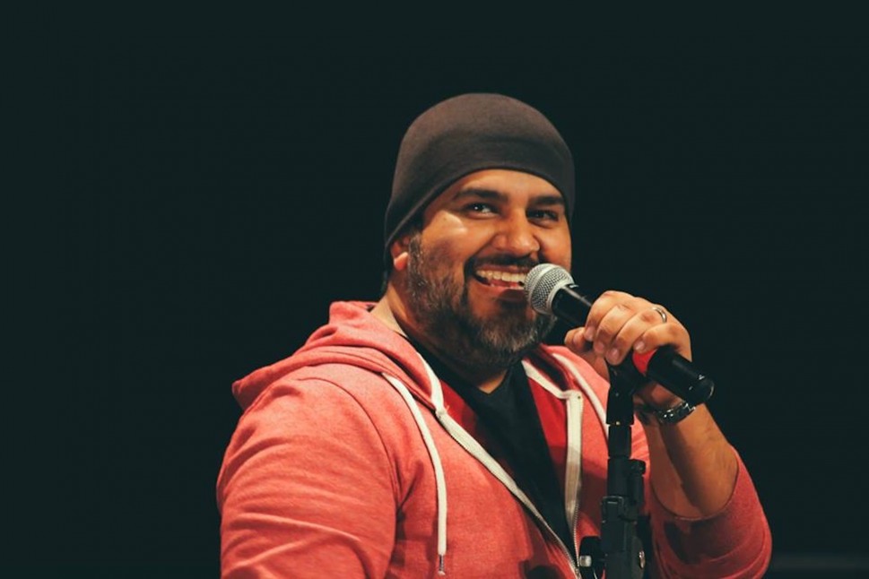Guadalupe Mendez, a Latino man, smiles as he performs at an open mic in an orange hoodie and black skullcap.