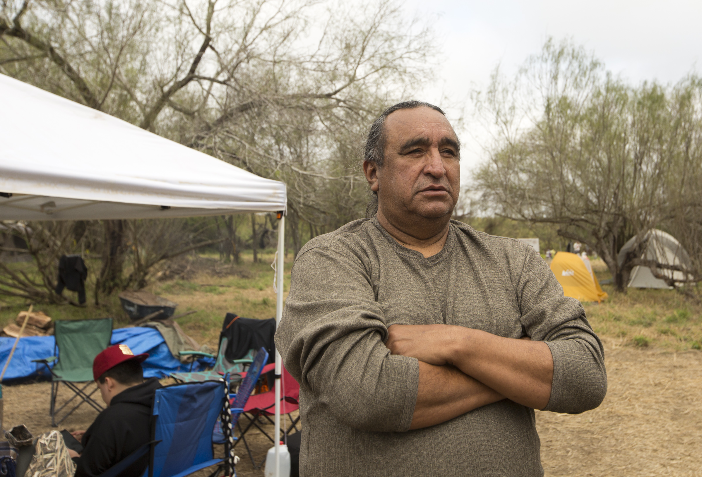 A Native American man in a gray long sleeve shirt stands with arms crossed and a distant, serious expression. Behind him are camp chairs with another tribesmember sitting in one.