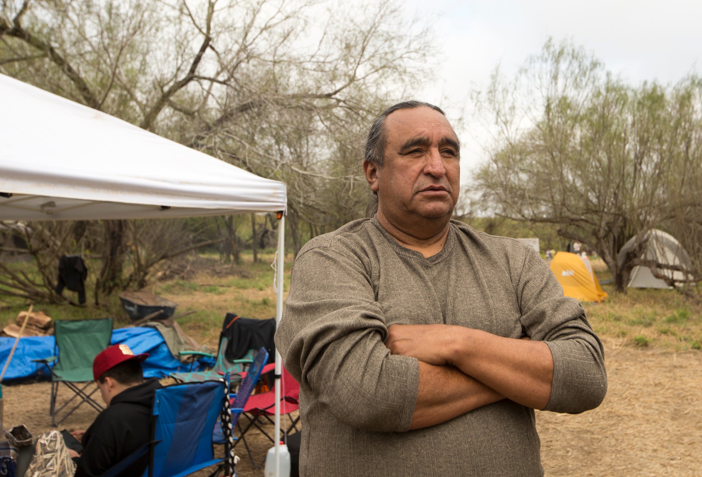 A Native American man in a gray long sleeve shirt stands with arms crossed and a distant, serious expression. Behind him are camp chairs with another tribesmember sitting in one.