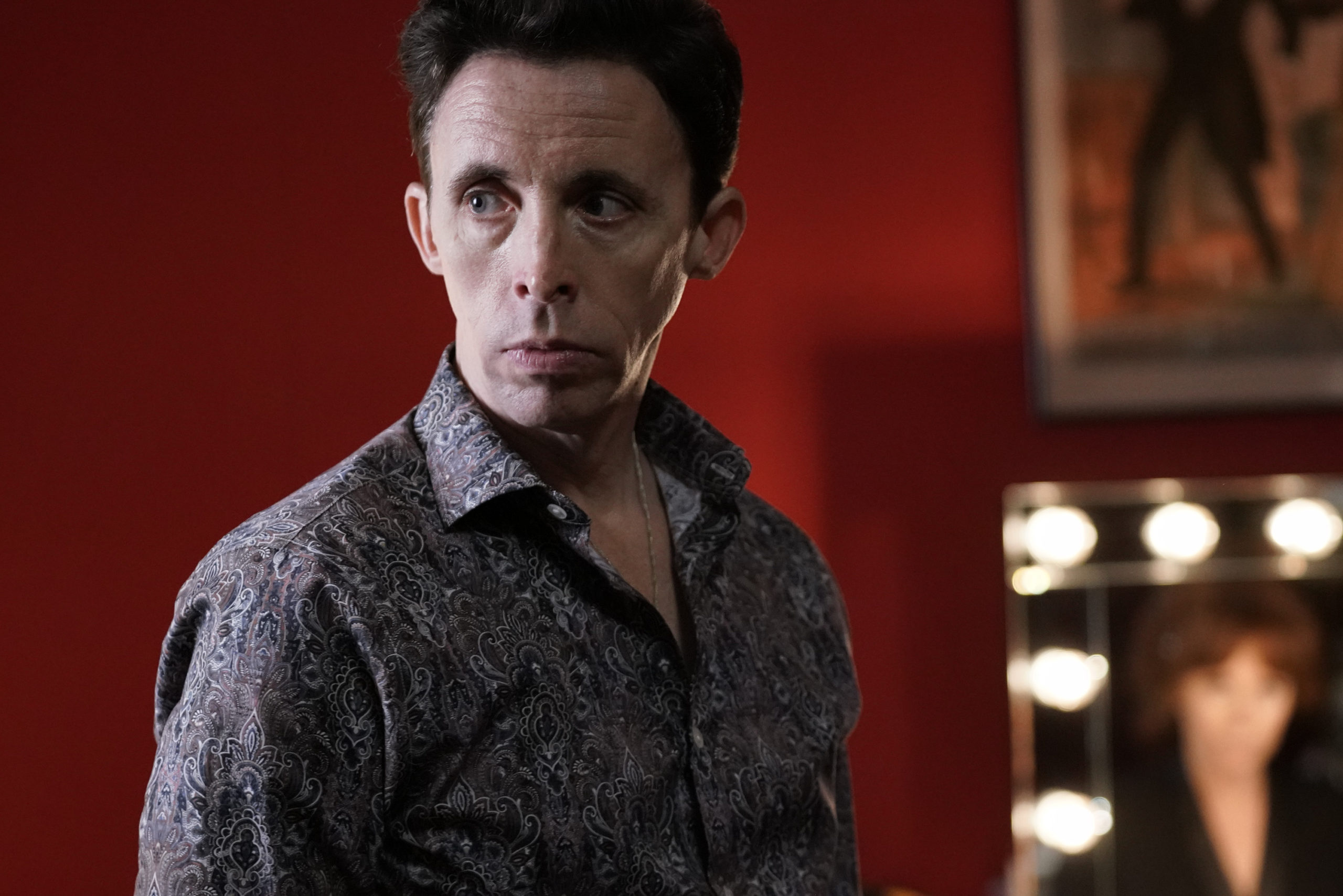 Kevin Cahoon, a white man with short brown hair, wears a worried expression and a dark colored, white-patterned button-down shirt. The lights of a dressing room mirror can be seen in the background.