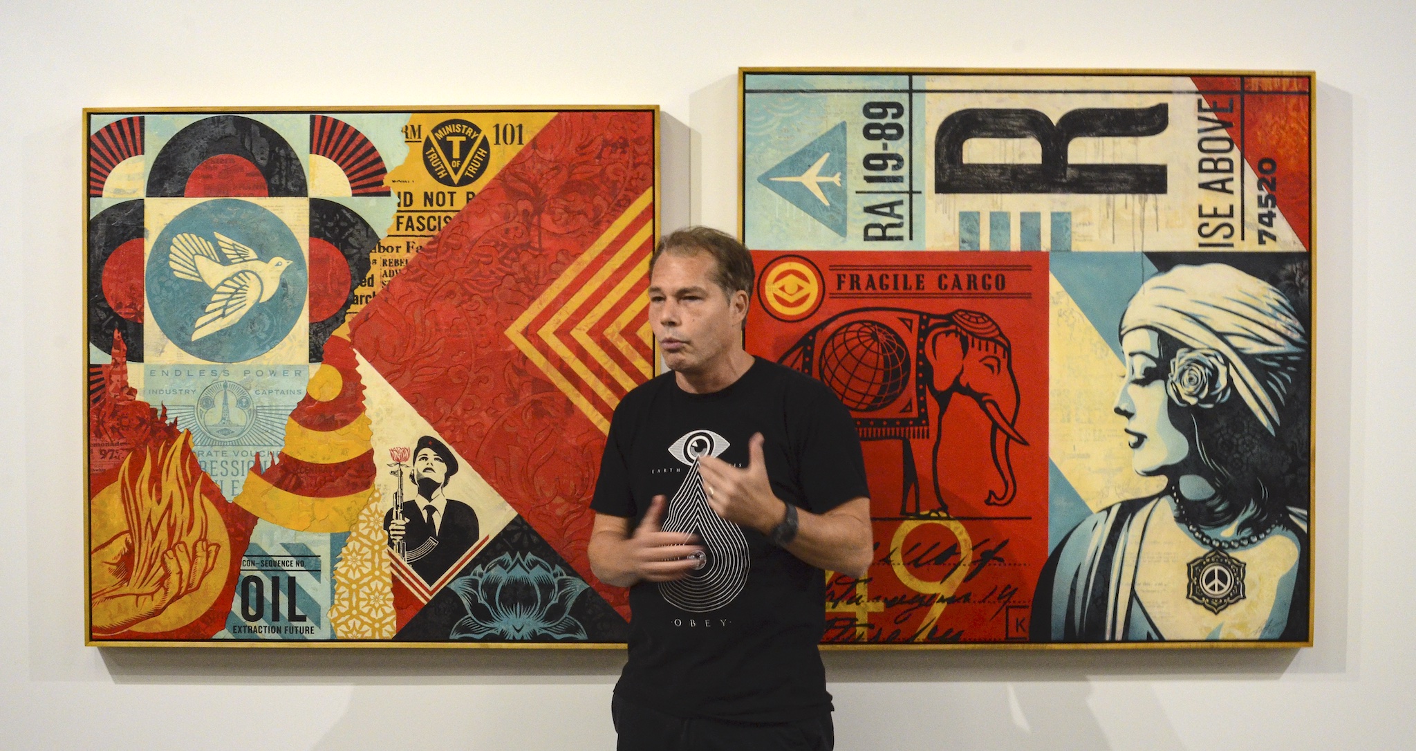 Shepard Fairey poses in front of two of his creations, colorful paintings dense with symbolism like guns, flowers, and an elephant. The overall look has a decoupage or collage effect.