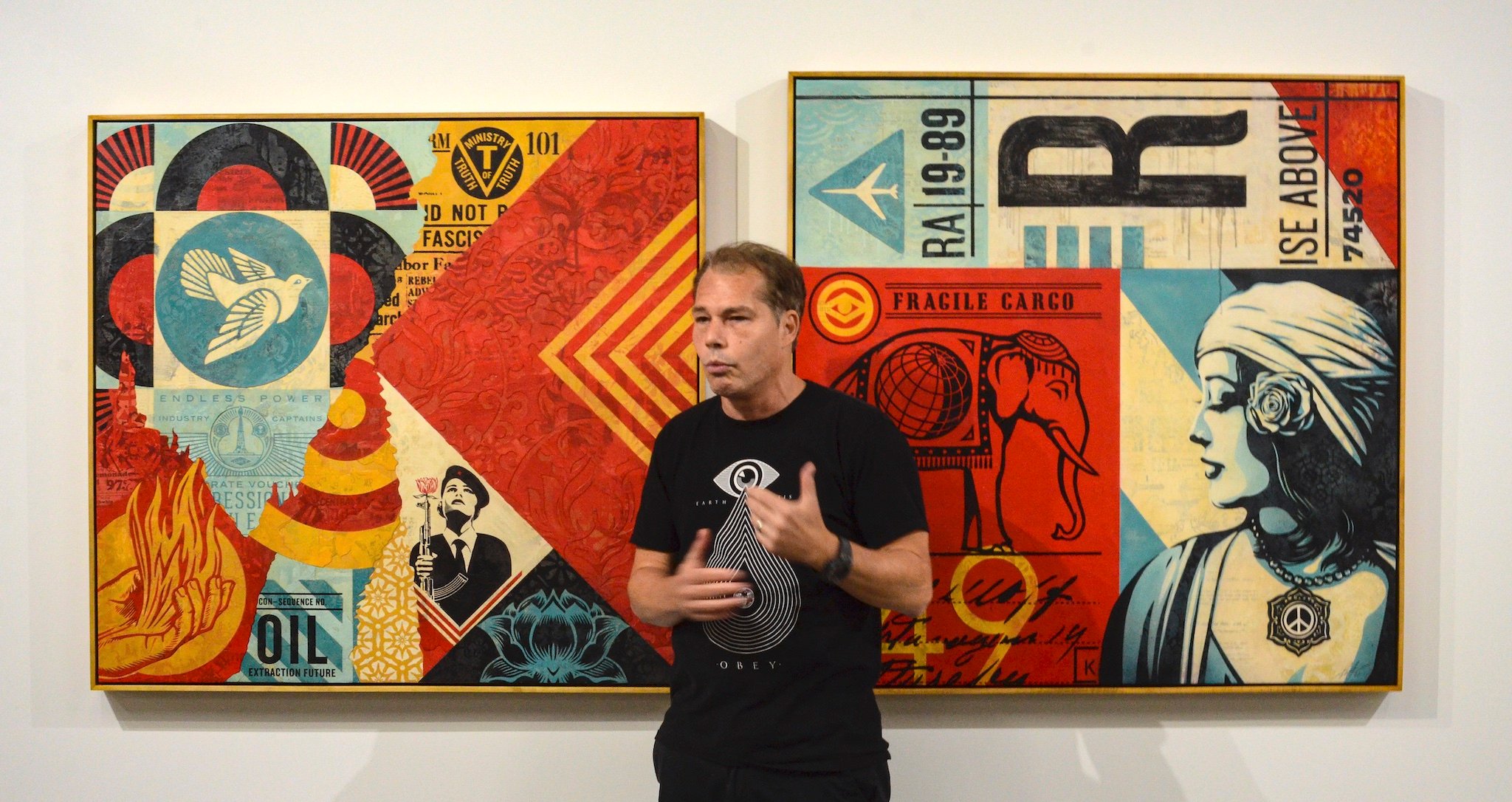 Shepard Fairey poses in front of two of his creations, colorful paintings dense with symbolism like guns, flowers, and an elephant. The overall look has a decoupage or collage effect.