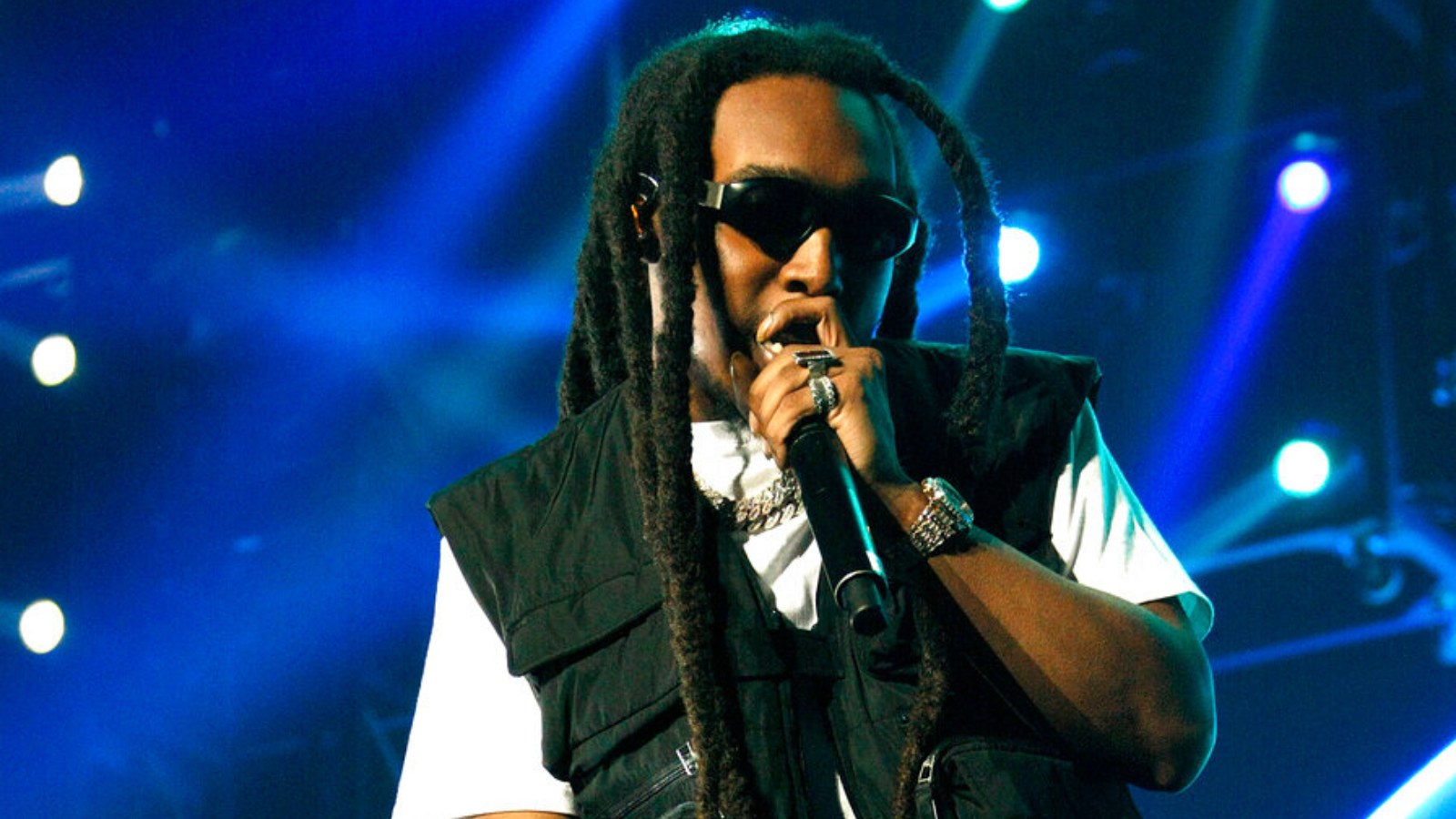 TakeOff, a Black man with thick dreaded braids, wearing sunglasses and a dark vest over a white shirt, raps into a microphone on stage.
