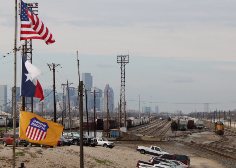 The Texas, United States and Union Pacific flags fly over the Englewood Union Pacific Rail Yard in Houston, which is full of trains, with the ground looking filthy between them.