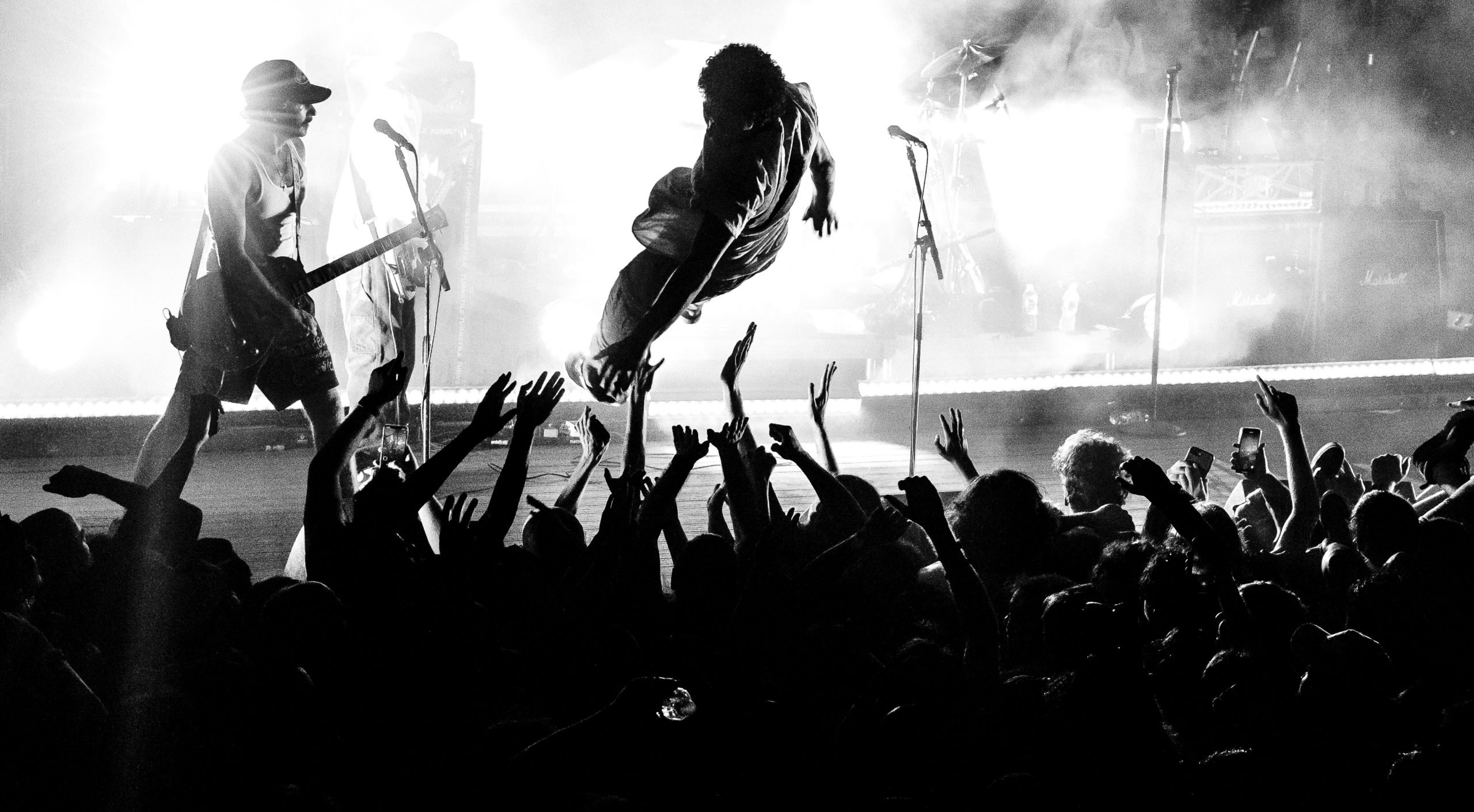 A black and white photo of a stage diver leaping into a crowd, who have their hands outstretched to catch the diver. In the background, a band plays hardcore music.