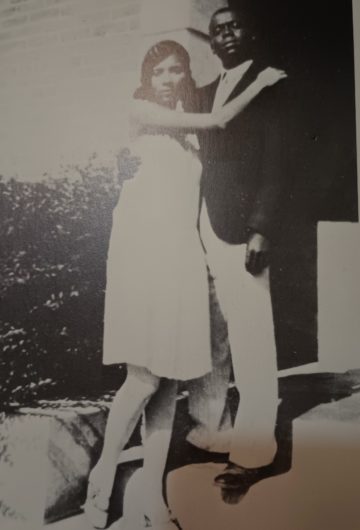 A faded photograph of Frank J. Robinson and Dorothy Redus Robinson in their younger days, with Dorothy embracing Fred as they stand on a stoop. Dorothy is in a white dress while Frank is wearing a jacket and slacks.