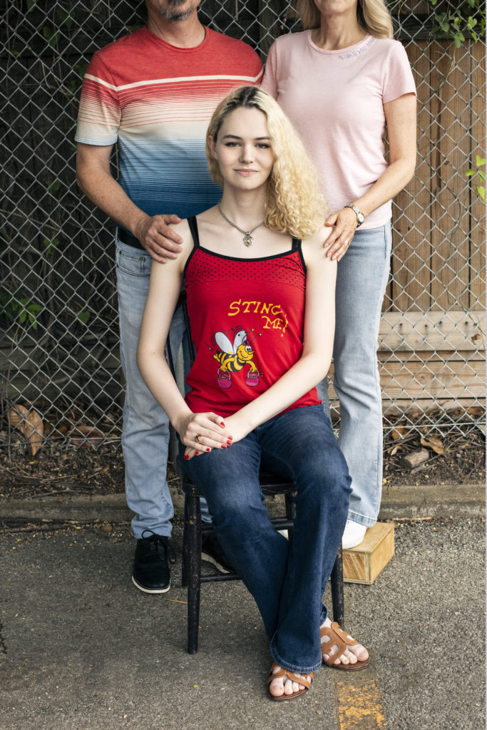 C smiles and looks directly at the camera. They have wavy blonde hair with dark roots showing and are wearing a red tank top with a cartoon bee on it, along with blue jeans. C's parents stand behind, each with a hand on C's shoulder.