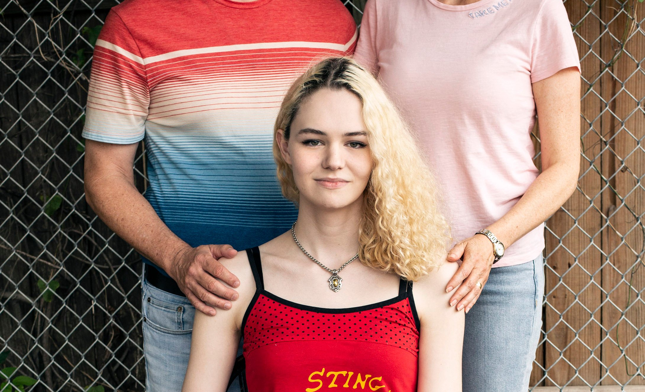 C smiles and looks directly at the camera. They have wavy blonde hair with dark roots showing and are wearing a red tank top with a cartoon bee on it, along with blue jeans. C's parents stand behind, each with a hand on C's shoulder.