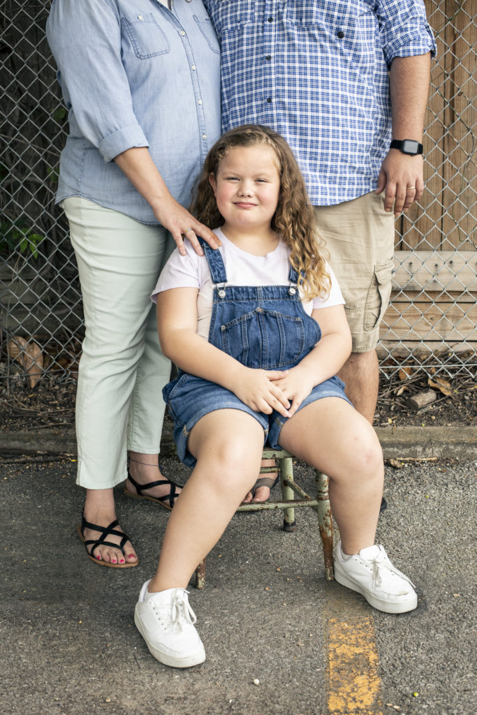 Cal is a young trans kid, wearing blue overalls, a white t-shirt, and white sneakers. Cal's parents stand behind, a hand on Cal's shoulder, dressed in blue button downs.