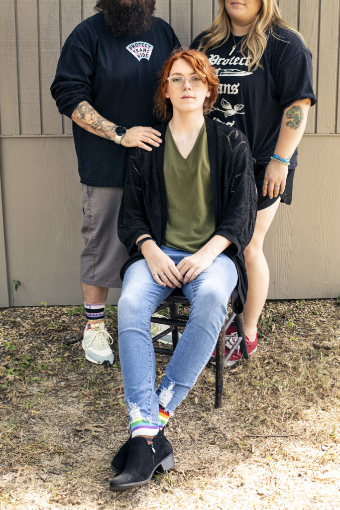 Jules looks directly at the camera. Jules has red hair, a green v-neck and black jacket along with blue jeans on. Their parents stand behind them wearing dark clothes, one with a hand on Jules' shoulder.