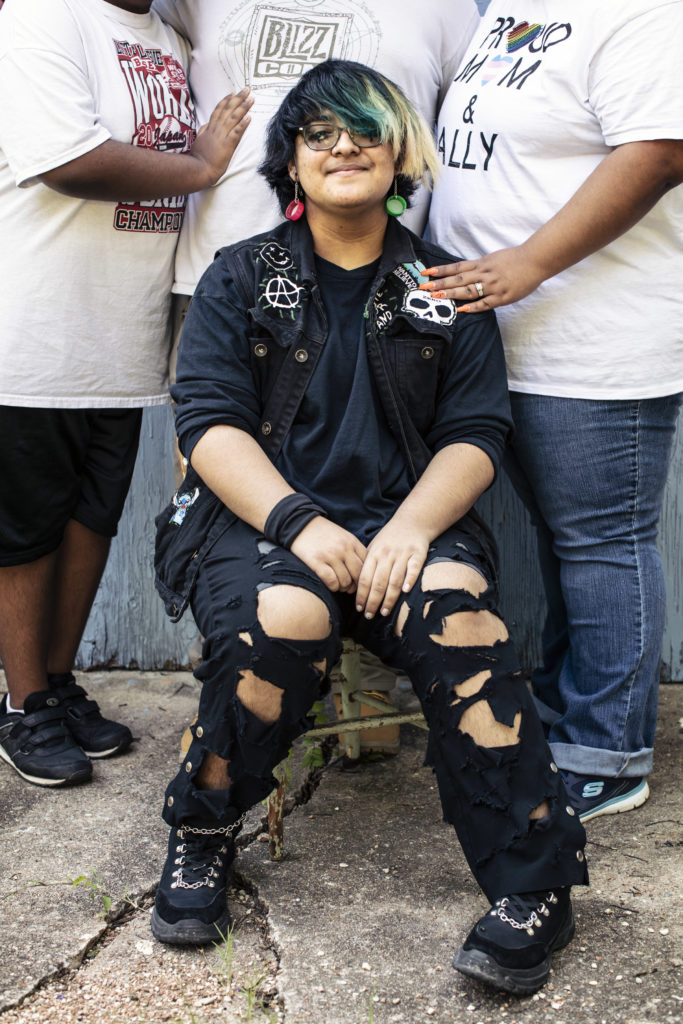 Justin has a very proud, happy expression as they look at the camera. They are wearing a punky black jacket with anarchy symbol and a skull, and torn black jeans, along with dark boots. Behind them are three adult family members, in white tees. One, with a hand on Justin's shoulder, has a tee reading "Proud Mom & Ally."