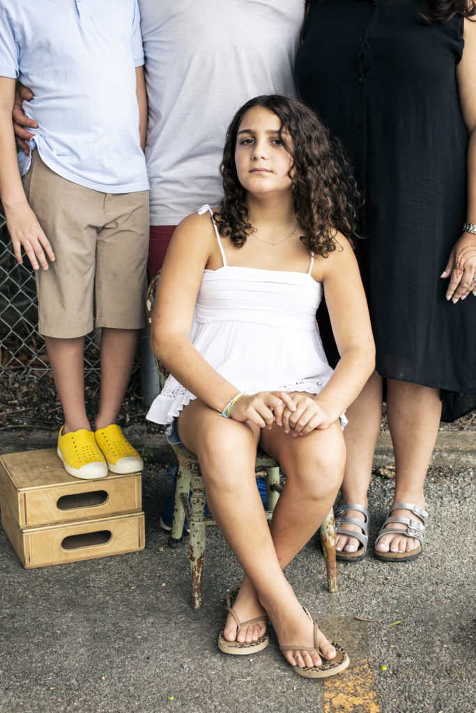 Maya is a young girl seated on a chair, with her parents and a sibling behind her, faces unseen. She has curly hair past her shoulders and she's wearing a simple white dress and sandals.