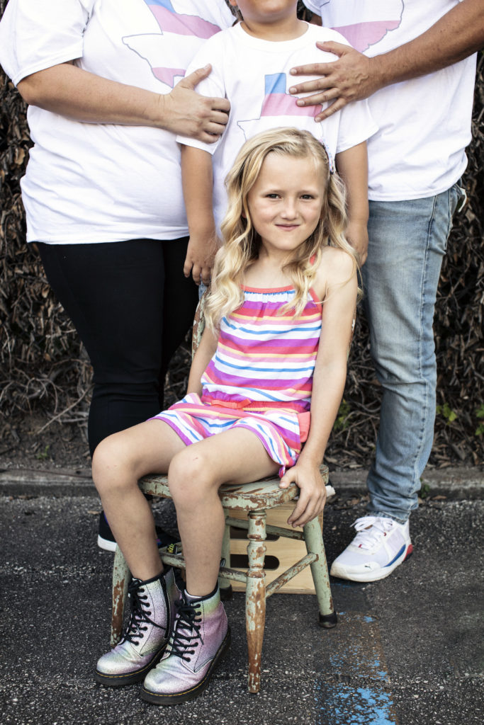 Sunny looks at us with a playful smile. Sunny's blonde hair spills over their shoulders, and they are wearing a pink and white dress and silver combat boots. Their family, including a sibling, stand behind them with faces out of sight.